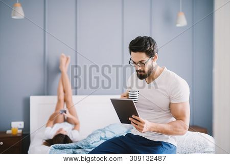 Attractive Muscular Man Drinking Coffee While Looking In A Tablet On The Bed In Front Of His Girlfri
