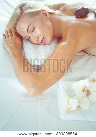 Beautiful Young Woman With Eyes Closed Receiving Hot Stone Massage At Salon Spa.