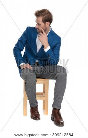 Concerned casual man looking down and thinking while holding his hand on chin and wearing a suit, sitting on white studio background