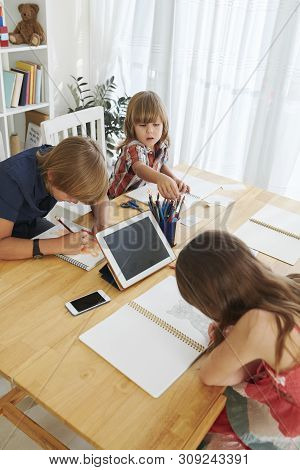 Group Of Children Drawing At Big Wooden Table With Gadgets, Pencils And Papep Cheets