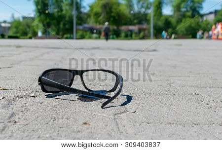 Broken Sun Glasses Lie On A Stone Tiled Walkway In The City Park Against The Background Of Residenti