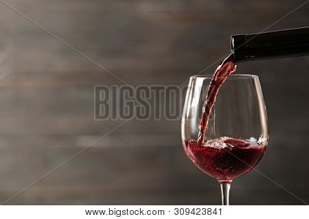 Pouring Red Wine Into Glass From Bottle Against Blurred Wooden Background, Closeup. Space For Text