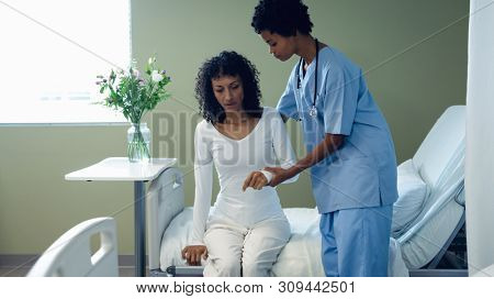 Front view of African-American female doctor helping mixed race female patient to get up on hospital bed in the ward at hospital