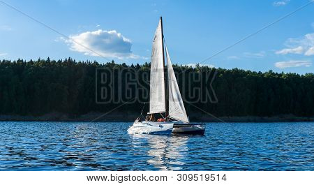People Rest On A Small Sailboat Catamaran Going Against The Wooded Shore
