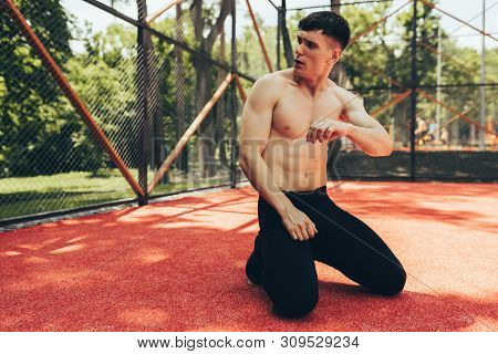 Handsome Fit Male Taking A Rest After Training Session Outdoors. Healthy Athlete Man Sitting At Pave