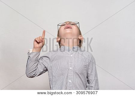 Portrait Of A Cute Blond Boy With Big Glasses. He Looks Up And Points A Finger. White Background.