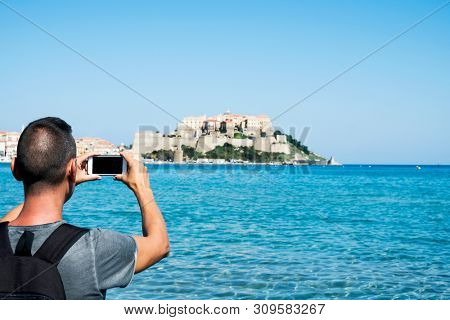 a young caucasian man, seen from behind, taking a picture of Calvi, in Corsica, France, with the Mediterranean sea in the foreground and the citadel of Calvi in the background