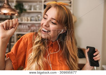 Image of a happy optimistic young blonde girl in orange shirt at the kitchen listening music with earphones.