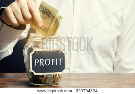 A Man Puts Dollars In A Glass Jar With The Word Profit. Concept Of Business Success, Financial Growt