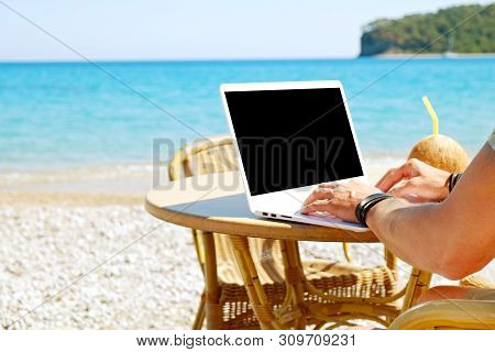 Young Man Working On His Laptop At The Beach At Tropical Destination. Office On The Beach Concept.