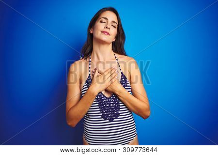 Beautiful woman wearing bikini swimwear over blue isolated background smiling with hands on chest with closed eyes and grateful gesture on face. Health concept.