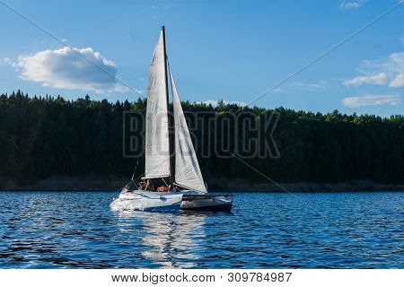 People Rest On A Small Sailboat Catamaran Going Against The Wooded Shore