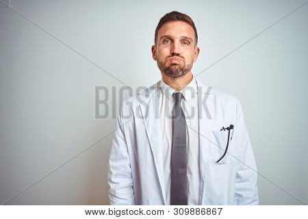 Young handsome doctor man wearing white profressional coat over isolated background puffing cheeks with funny face. Mouth inflated with air, crazy expression.