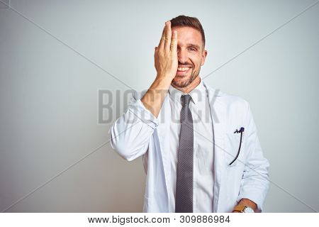 Young handsome doctor man wearing white profressional coat over isolated background covering one eye with hand, confident smile on face and surprise emotion.