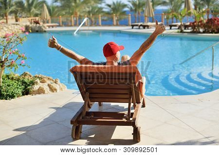 Relax In Summer Pool. Young And Successful Man Lying On A Sun Lounger And Raising Arms At The Hotel 