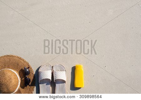 Summer Vacation Concept: Beach Slippers, Protective Cream And A Hat On Clean Sand. Top View Of Acess