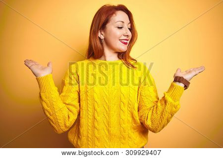 Beautiful redhead woman wearing winter sweater standing over isolated yellow background smiling showing both hands open palms, presenting and advertising comparison and balance