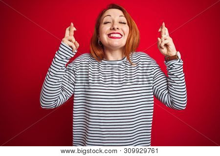 Young redhead woman wearing strapes navy shirt standing over red isolated background gesturing finger crossed smiling with hope and eyes closed. Luck and superstitious concept.