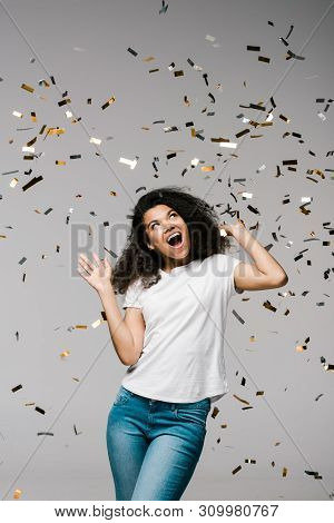 Young African American Woman Smiling And Looking At Falling Confetti While Standing On Grey