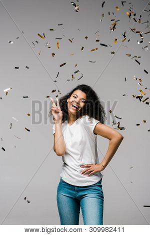 Young African American Woman Smiling Near Shiny Confetti While Standing With Hand On Hip And Showing