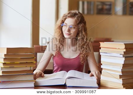 Young European Sweet Curly Haired Girl Looking Aside, Sitting At Desk. Spending Her Time In Library,