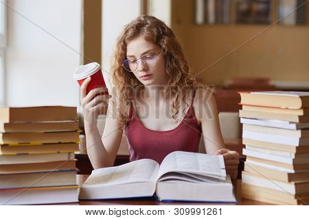 Attracvtive Young Lady Wearing Glasses And Casual Uotfit, Reading Book Which Laying On Table And Hol