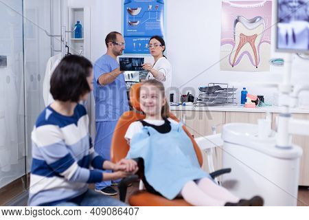 Dentist And Assistant In Dental Office Holding Little Girl Jaw Radiograph Discussing Diagnosis.. Chi