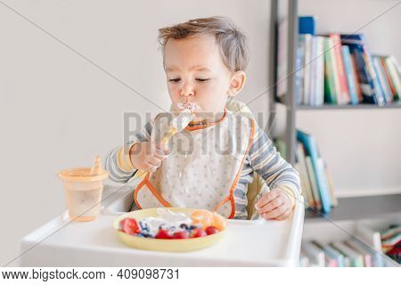 Cute Caucasian Baby Boy Eating Ripe Berries And Fruits With Yogurt. Funny Smiling Child Kid Sitting 
