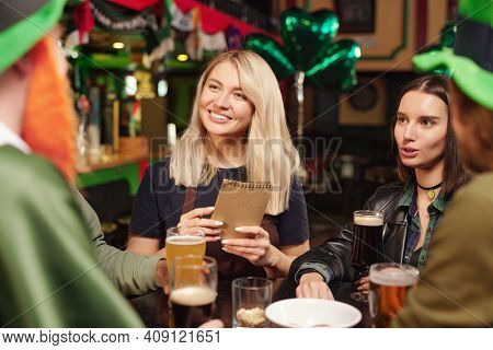 Young women sitting at the table talking to friends and drinking beer during celebration of Patricks Day in the bar