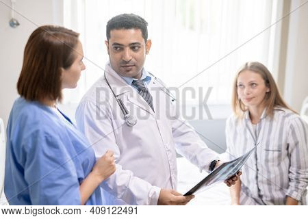 Young sick woman sitting on bed and looking at confident doctor discussing x-ray results with his assistant in hospital chamber