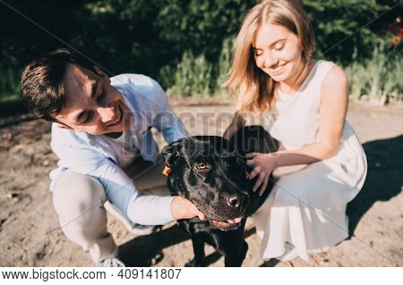 Lovely Couple Is Walking With A Black Labrodor On A Leash. Lovers Smile And Watch The Dog. A Walk In