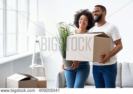 Loving Black Couple Holding Packed Boxes Moving House, Posing Standing Together In New Apartment Ind