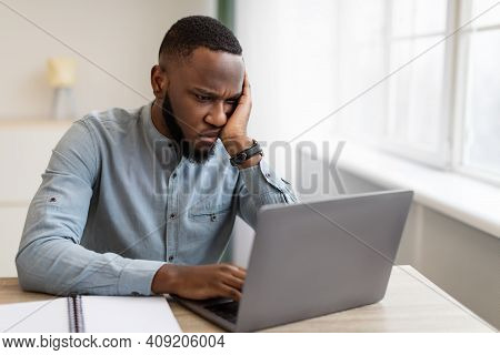 Unhappy Black Businessman Looking At Laptop Hating His Boring Job Sitting At Workplace In Modern Off
