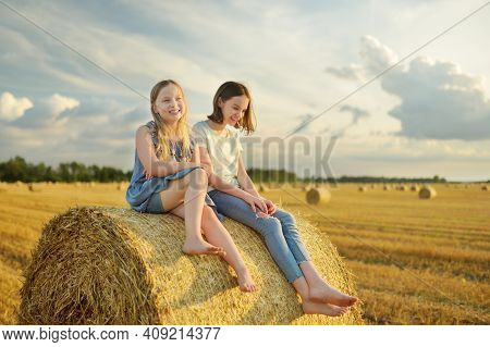 Adorable Young Sisters Having Fun In A Wheat Field On A Summer Day. Children Playing At Hay Bale Fie