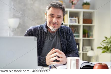 Bearded man working online with laptop computer at home sitting at desk. Home office, browsing internet, study room. Portrait of mature age, middle age, mid adult man in 50s.