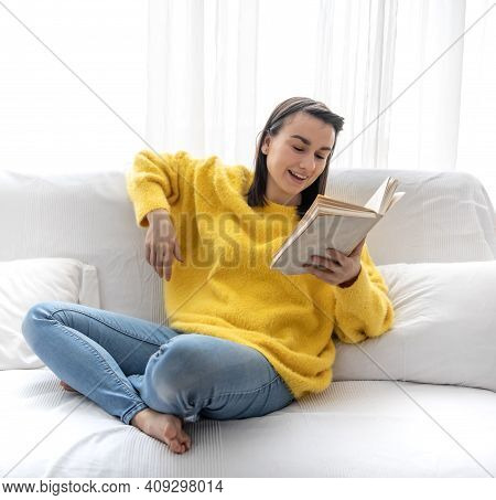 Stylish Girl In A Yellow Sweater Is Resting At Home On The Sofa With A Book.