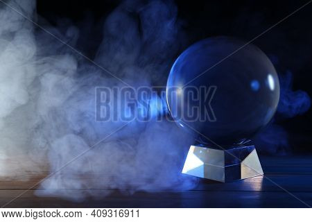 Magic Crystal Ball On Wooden Table And Smoke Against Dark Background, Space For Text. Making Predict