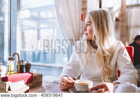 Blonde Middle Age Woman Sit Alone In Cafe With Cup Of Coffee And Look To Window