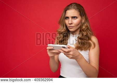 Serious Beautiful Young Blonde Woman Wearing White T-shirt Isolated Over Red Background Using Smartp