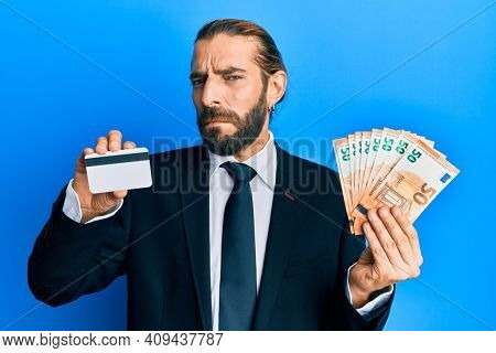 Attractive business man with long hair and beard holding credit card and euros in shock face, looking skeptical and sarcastic, surprised with open mouth 