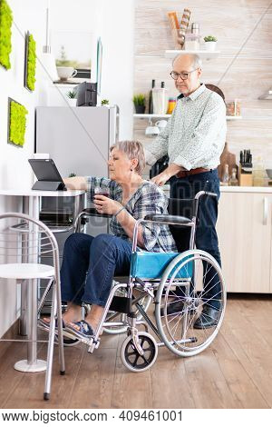 Disabled Senior Woman In Wheelchair Using Tablet Computer In Kitchen With Husband Near. Paralysied H