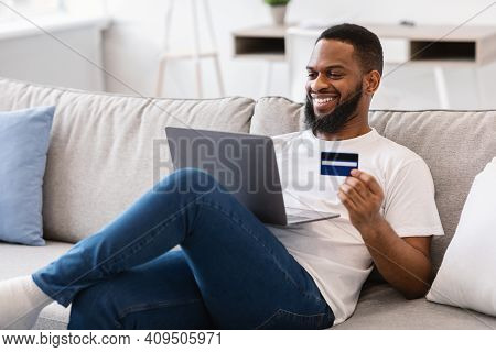 African Guy Holding Credit Card Shopping Online Via Laptop Computer Sitting On Couch At Home. Happy 