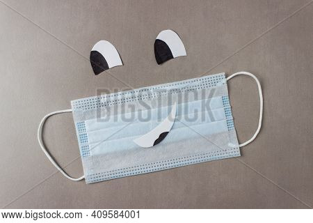 Blue Disposable Medical Mask With A Drawn Face - Smile And Eyes On A Gray Background.