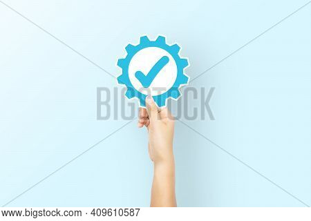 Hand Holding Paper With Icon Gear And Check Mark On Light Blue Background. Concept Of Standard Quali