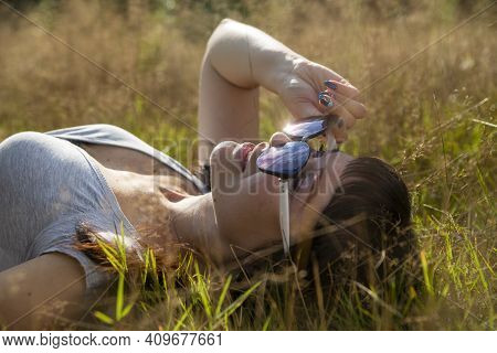 Happy Beautiful Young Woman In Sunglasses Smiles And Lies On A Meadow In The Grass In The Sunshine E