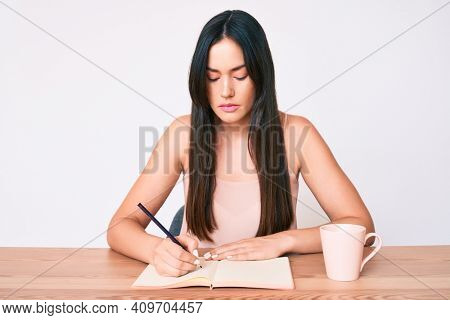 Young caucasian woman sitting at the desk writing book drinking coffee thinking attitude and sober expression looking self confident 