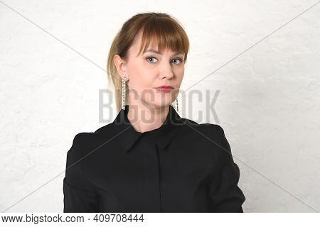 Young Woman With Hair Pulled Back And Bangs In Black Dress With Round Collar Against White Wall. Min