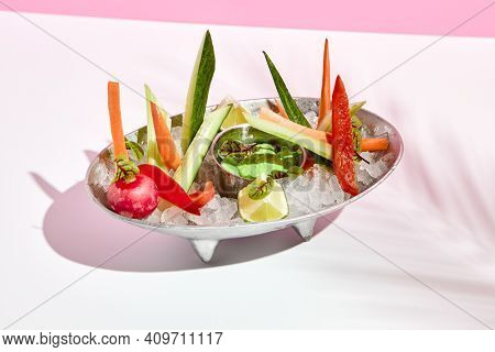 Veggie plate with crushed ice and green dip. Vegan appetizer plate on white table with pink wall. Day sunlight with hard shadow of fern palm leaves. Summer or spring restaurant food concept