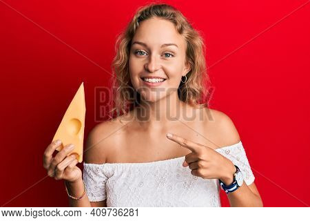 Beautiful caucasian woman holding a piece of emmental cheese smiling happy pointing with hand and finger 