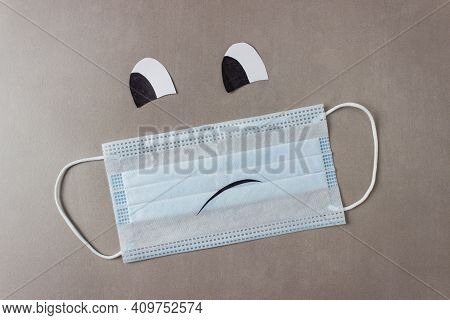 Blue Disposable Medical Mask On A Gray Background With Sad Smiley Face With Drawn Eyes And Mouth